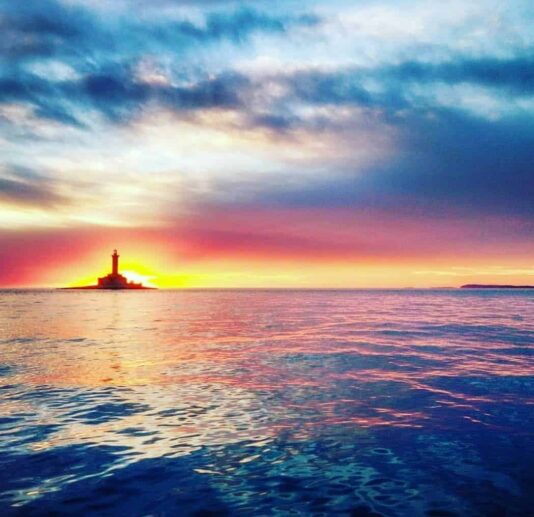 A vibrant sunset over the Adriatic Sea with a silhouette of a lighthouse in the distance.