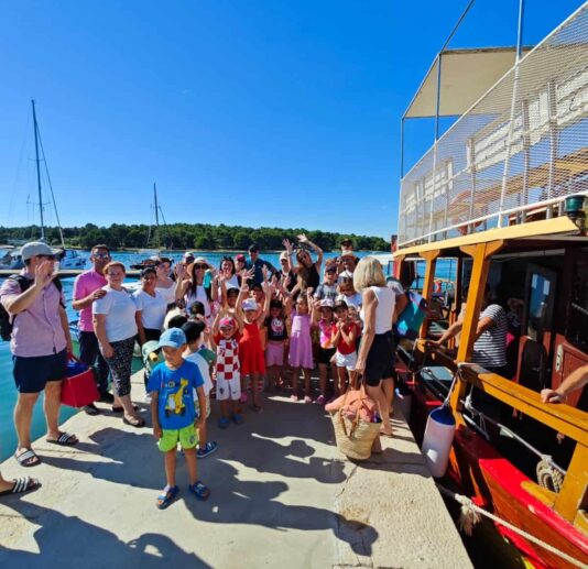 A group of smiling people, including children, gathered by a colorful boat at the Medulin harbor.